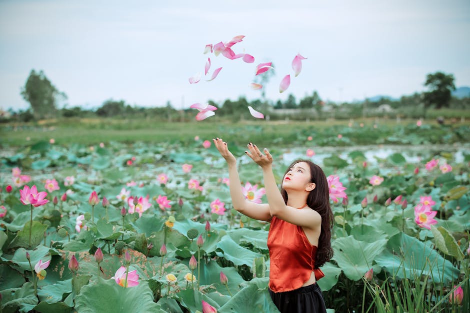 An adult woman in a vibrant lotus garden enjoying falling petals on a sunny day.