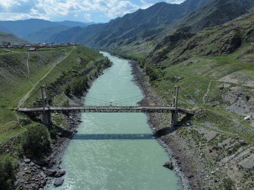 Free A picturesque bridge crossing a serene river with mountainous landscape and cloudy sky. Stock Photo
