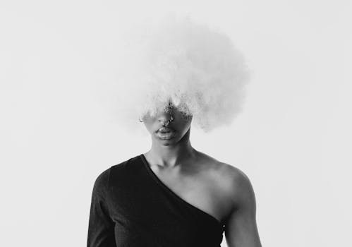 Artistic black and white portrait of a woman wearing a large afro wig in a minimalistic studio setting.