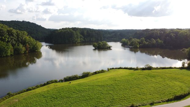 A tranquil aerial view of a picturesque lake surrounded by lush green trees and rolling hills on a sunny day.