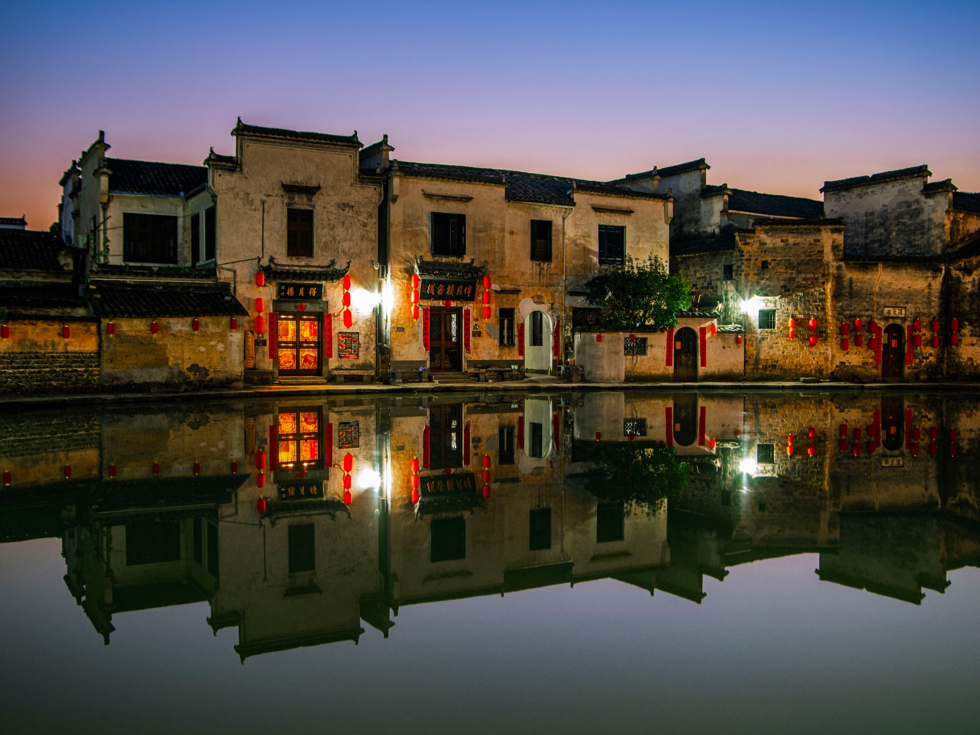 Charming view of traditional Chinese buildings and lanterns reflecting in water at twilight.
