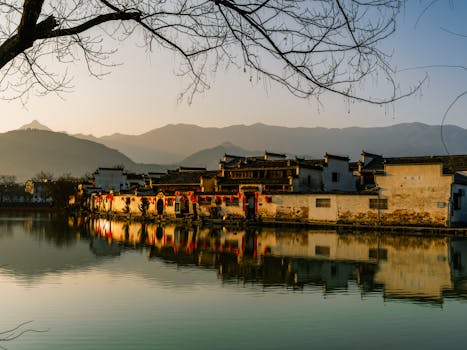 A tranquil landscape of an Asian village with calm waters and mountain backdrop, captured at sunset.