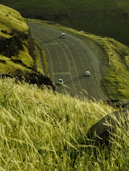 A scenic high-altitude road winding through lush green hills, featuring cars driving through a serene natural landscape.