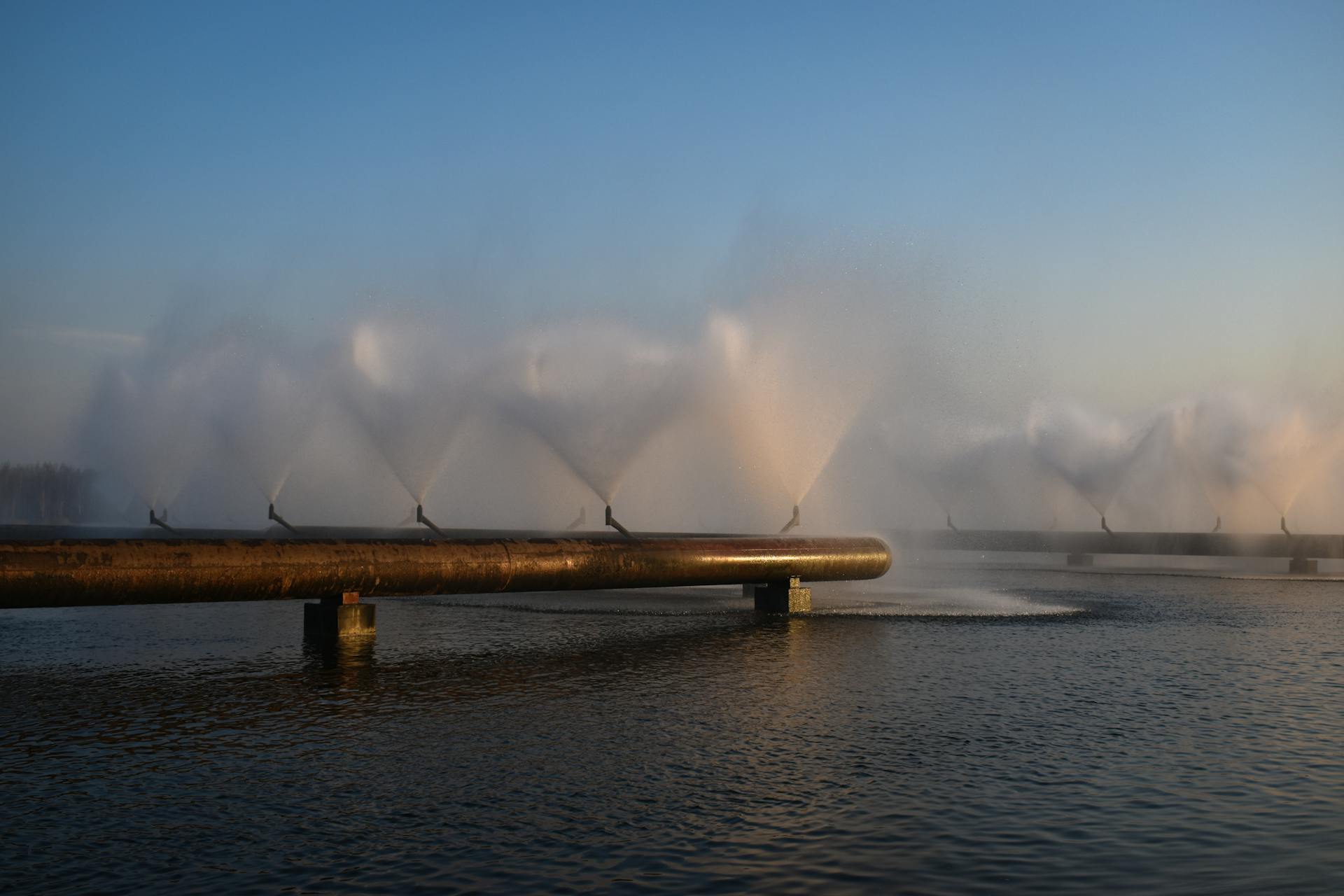 A scenic view of industrial water pipes creating fountain effects over a calm water surface at sunset.