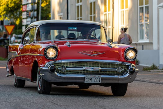 A vintage red Chevrolet drives down a sunlit urban street, showcasing classic design and style.