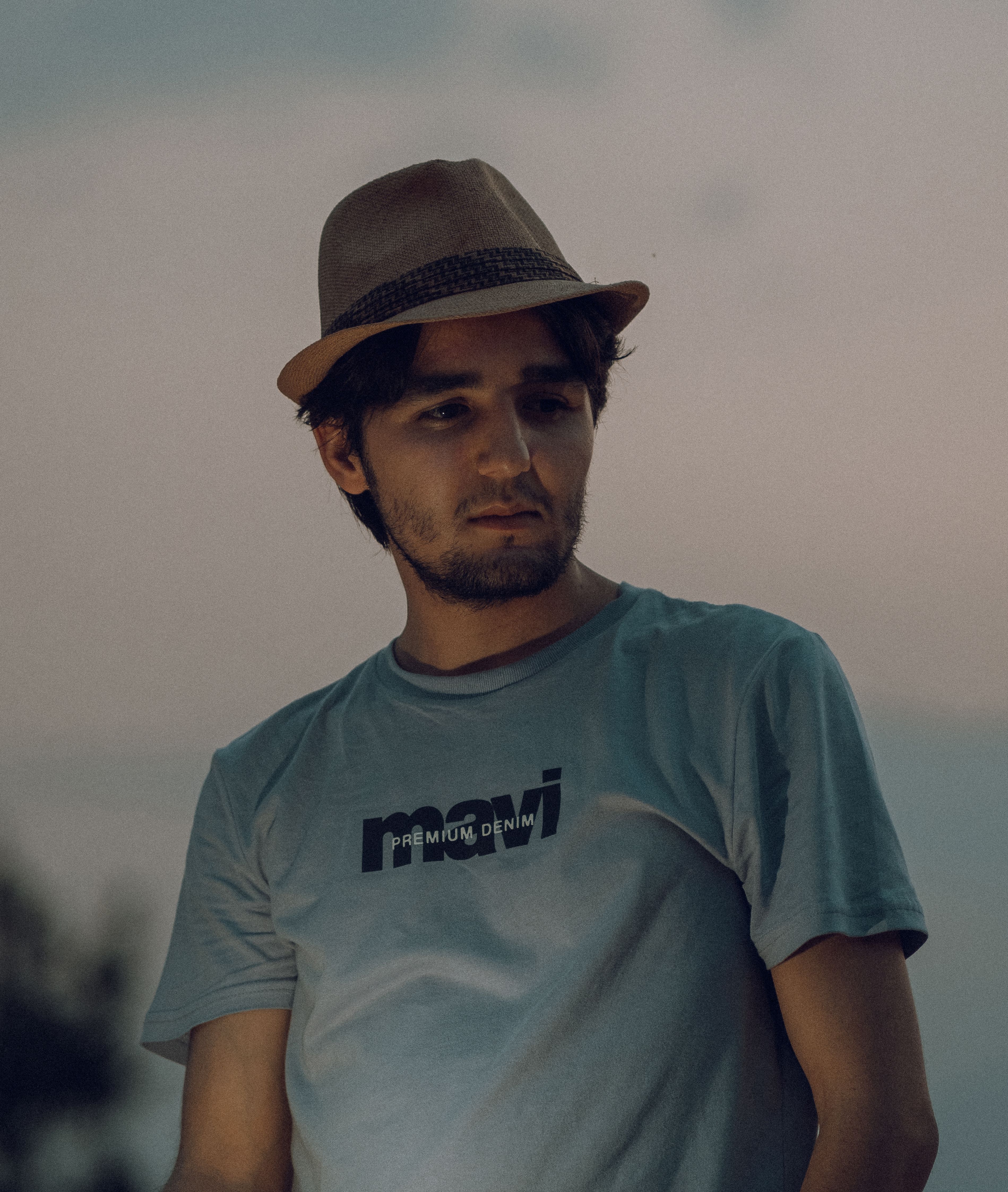 A contemplative man wearing a fedora and a casual shirt, captured at twilight.