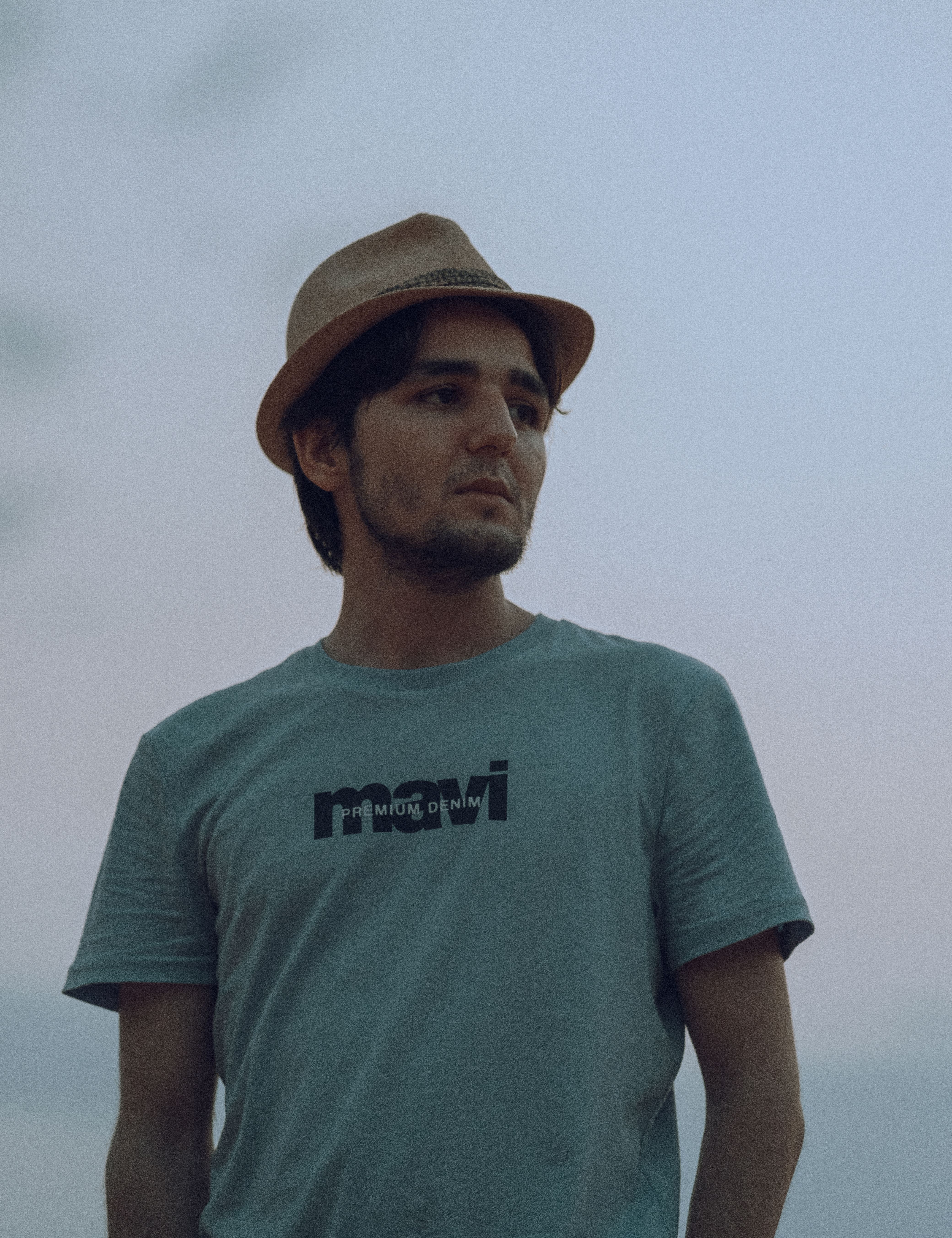 Casually dressed man wearing a hat, looking thoughtful under a clear sky.