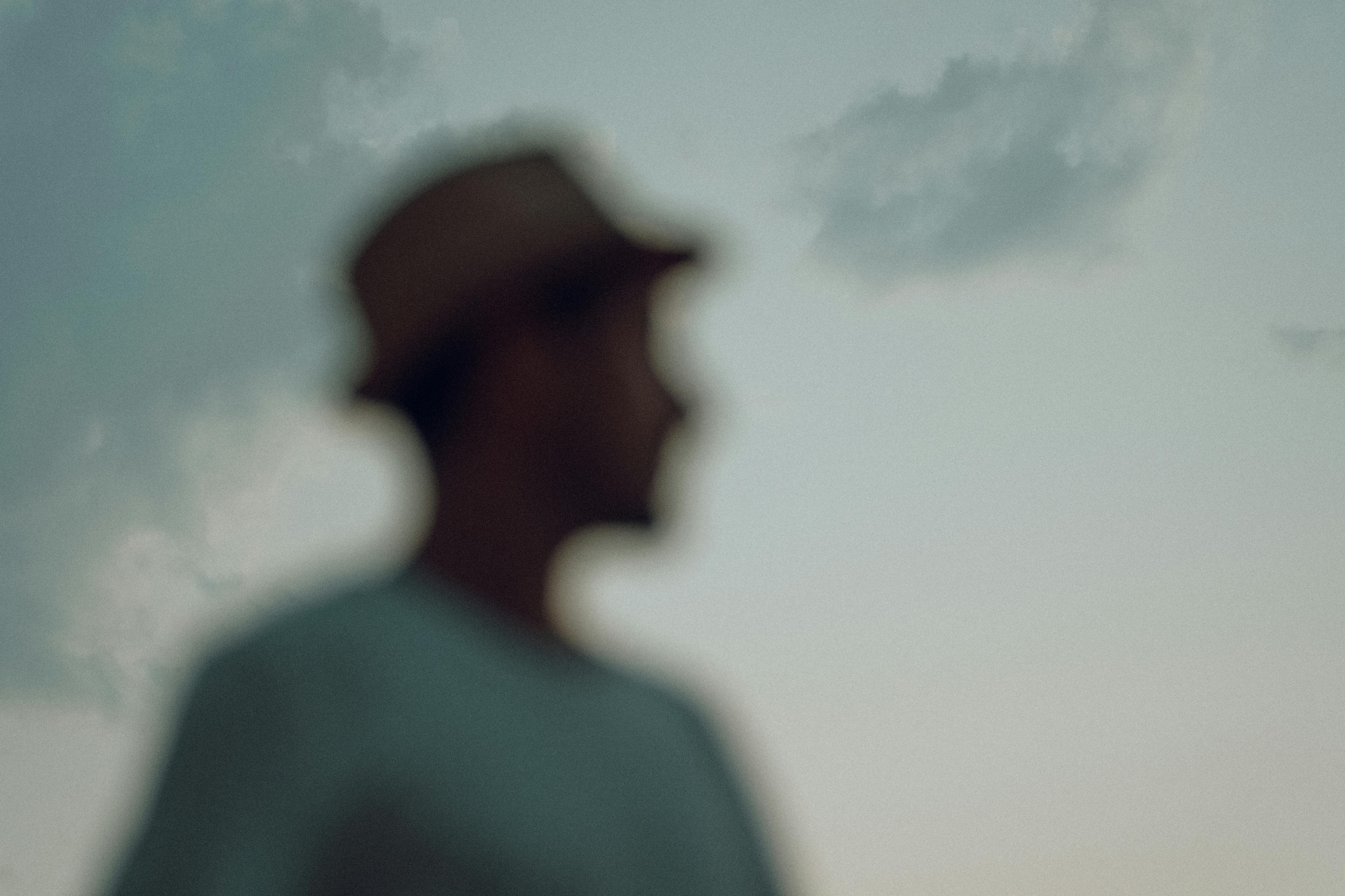A blurred silhouette of a man wearing a hat, captured during twilight with a serene sky as background.