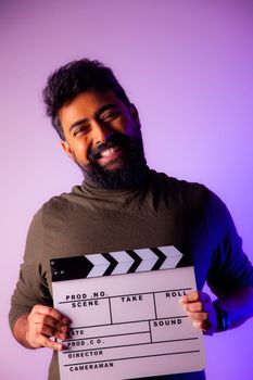 Portrait of a smiling bearded man with a clapperboard against a colorful backdrop.