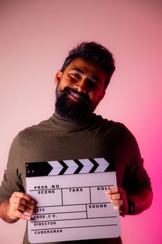 Smiling man holding a film slate against a pink background in a studio setting