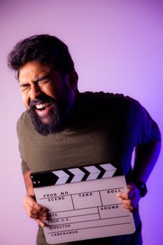 A bearded man laughs while holding a clapperboard during a studio shoot with dramatic lighting.
