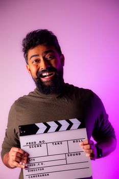 Portrait of a cheerful bearded man with a film slate, set against a vibrant purple background.
