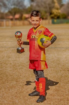 A young boy in a red jersey holding a trophy and soccer ball on a sunny field.
