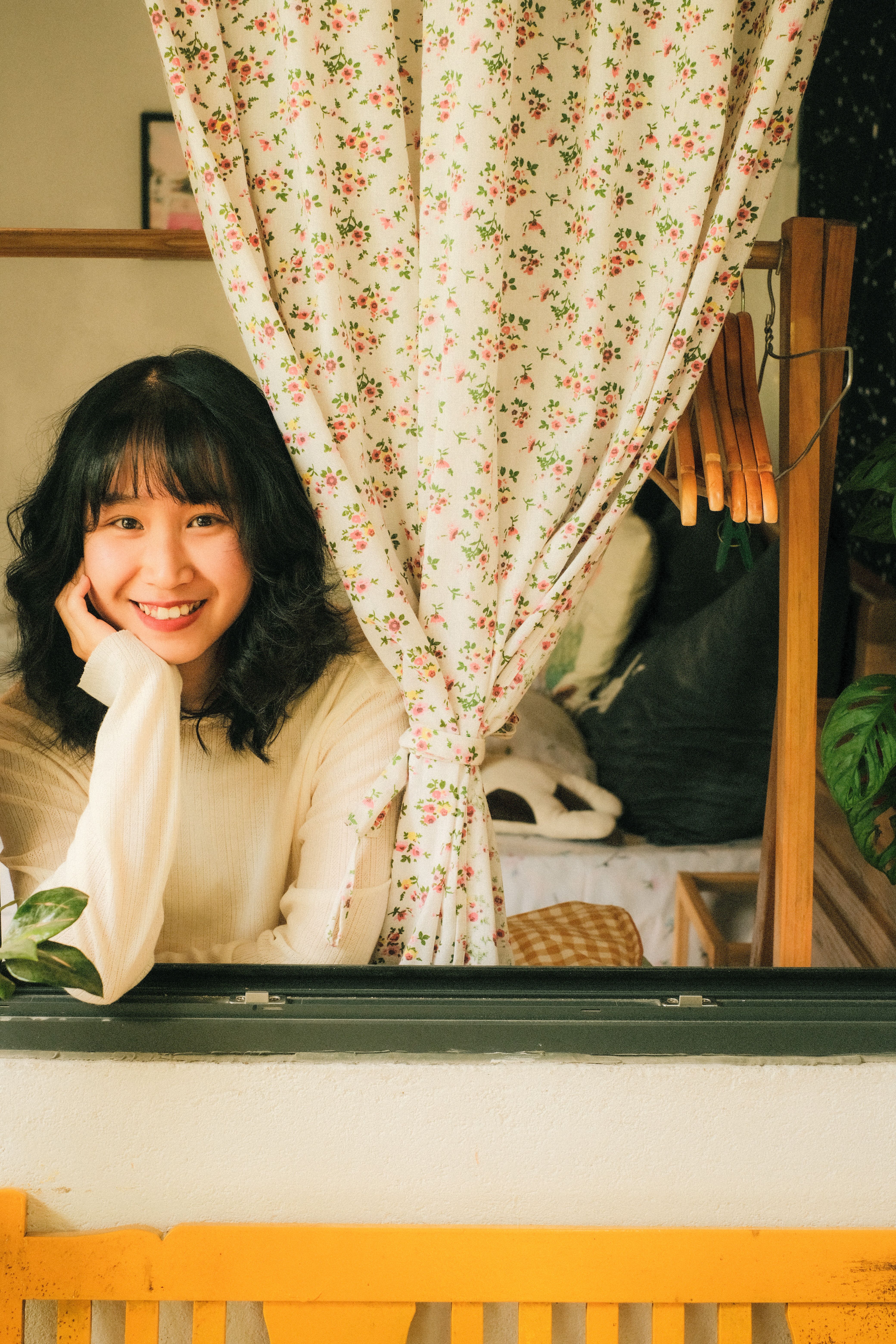 Asian woman smiling happily beside a floral curtain, exuding warmth and charm.