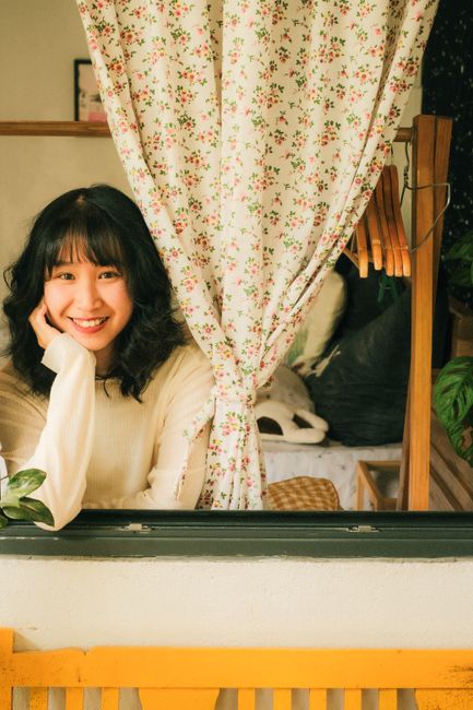 Asian woman smiling happily beside a floral curtain, exuding warmth and charm.
