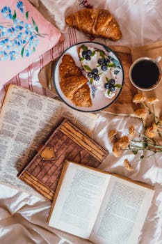 Top view of a cozy breakfast scene with croissants, coffee, and books on a bed.