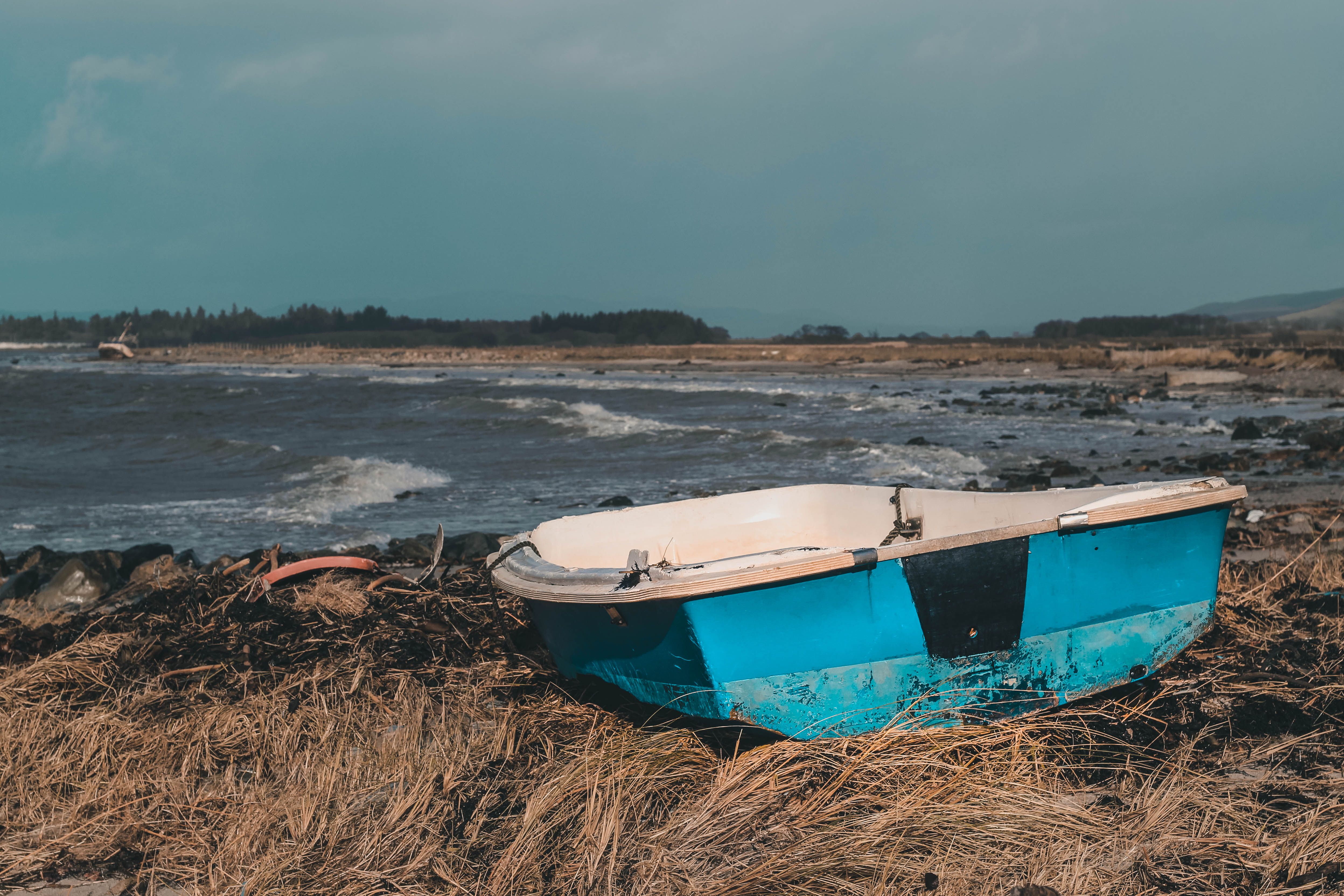 Blue boat on a grassy shore in Campbeltown, Scotland, with waves and distant hills.