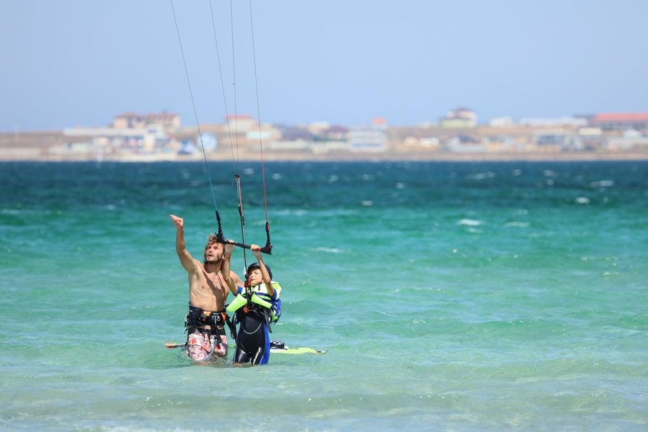 A young boy learns kiteboarding from an adult in the clear sea near a town.