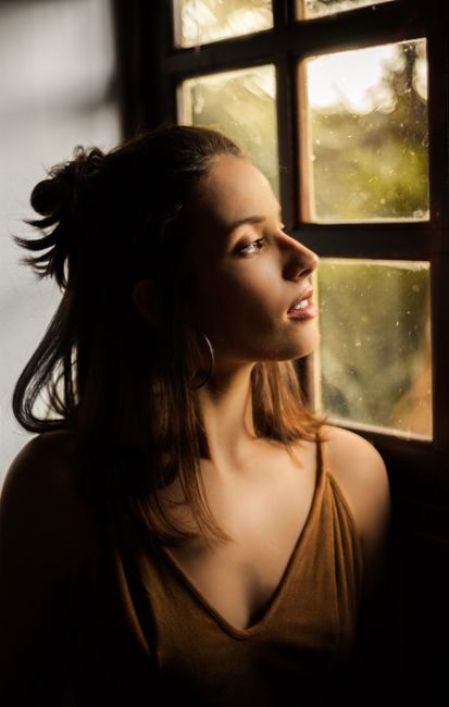 A woman gazes thoughtfully through a window during a warm golden hour.