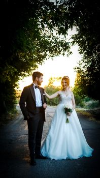 Bride and groom standing on a tree-lined road during sunset, sharing a joyful moment.