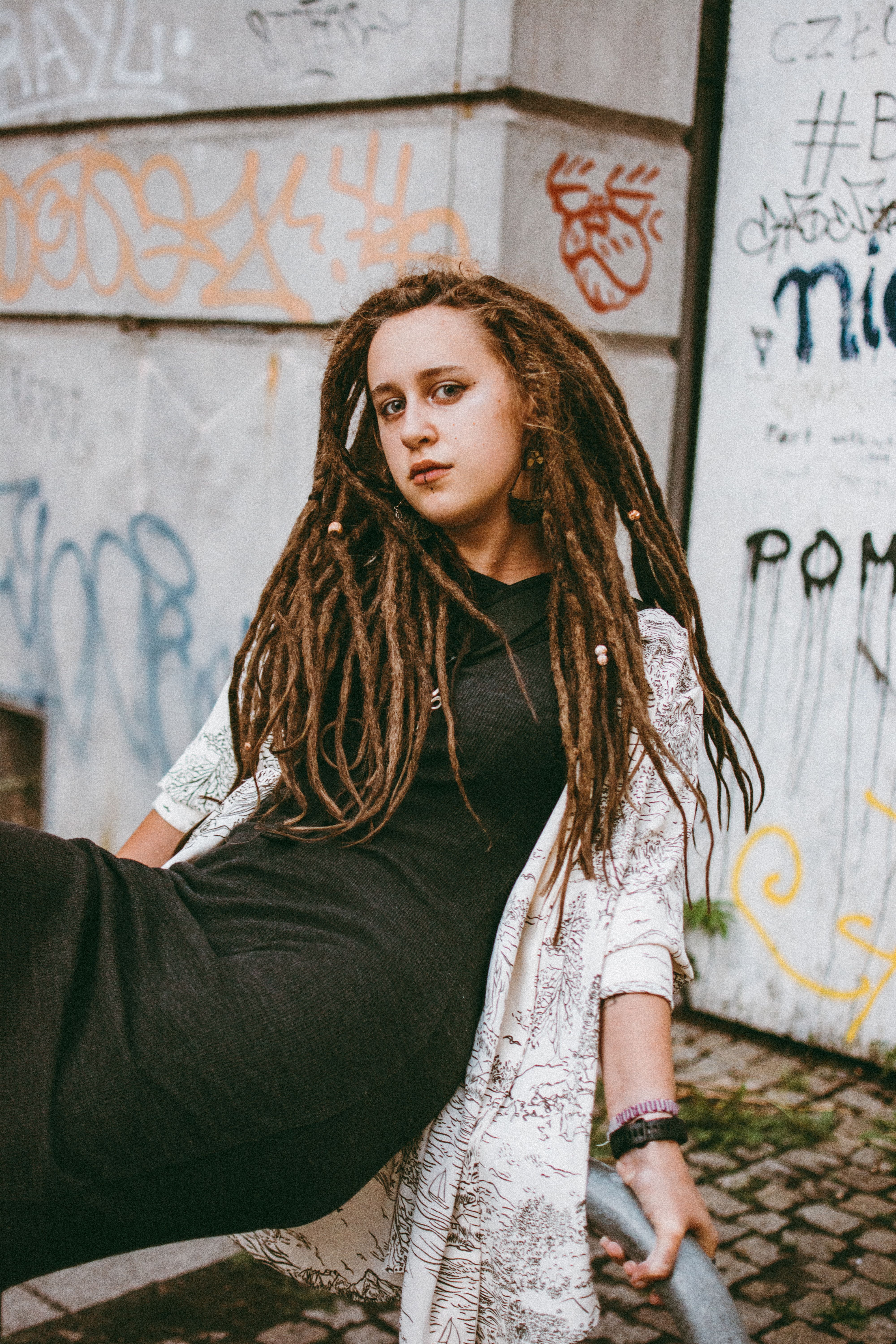 Woman with dreadlocks posing confidently against a graffiti wall in an urban environment.