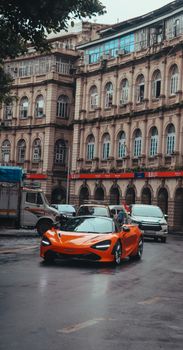 An orange sports car drives through a historic city street under overcast skies.