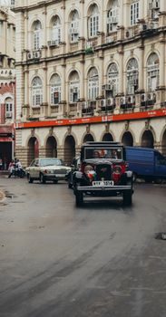 A classic vintage car driving in front of a historic building in a bustling city.