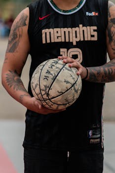 Close-up of a tattooed athlete holding a worn basketball, wearing a Memphis jersey.