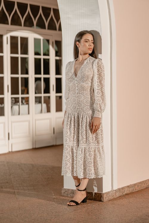 Free A woman in a white lace dress stands elegantly against an arched wall indoors. Stock Photo