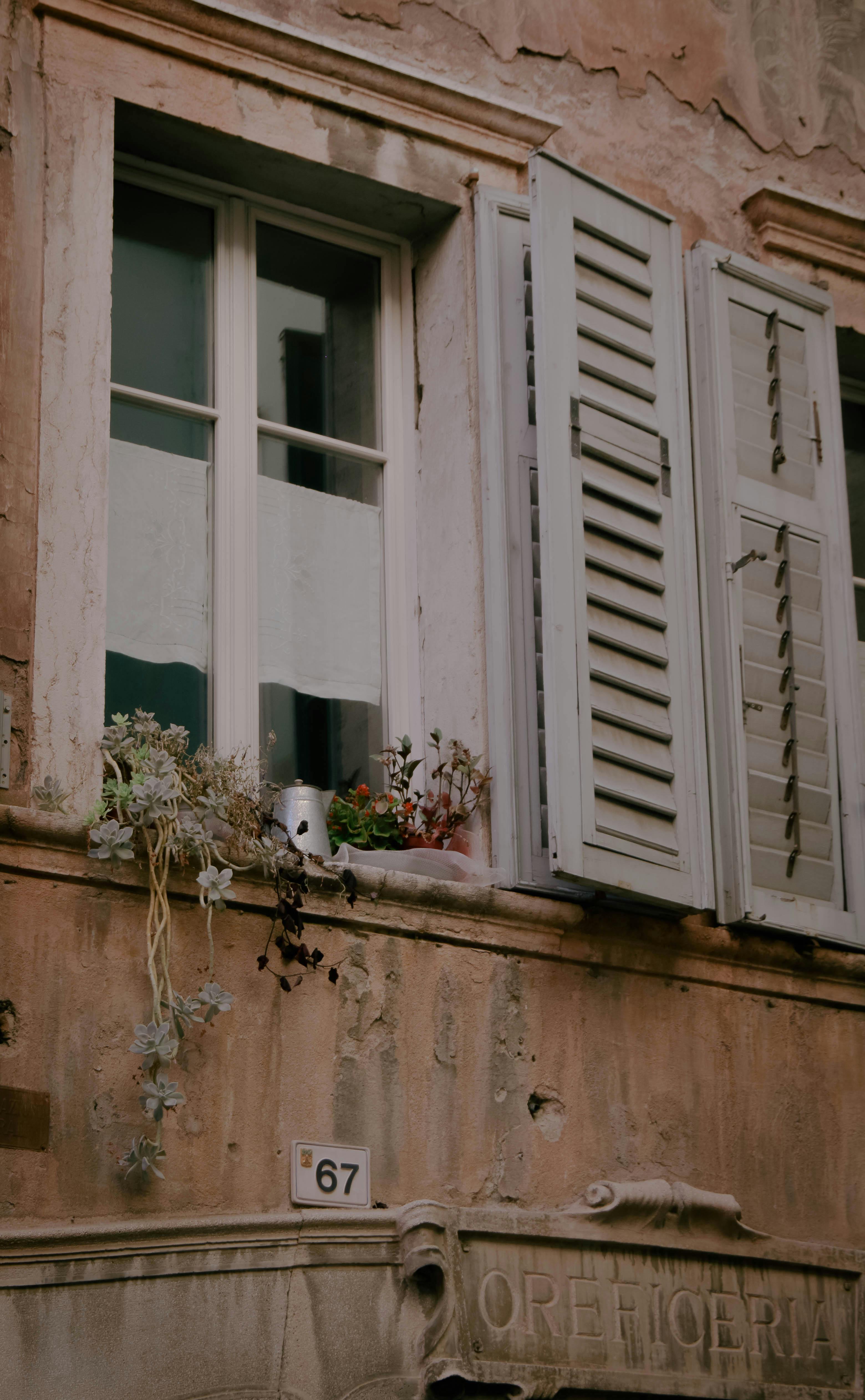 A rustic wall with open shutters and potted plants in Rovereto, Italy.