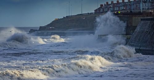 Free Powerful ocean waves crash against a coastal wall near residential buildings, under a cloudy sky. Stock Photo