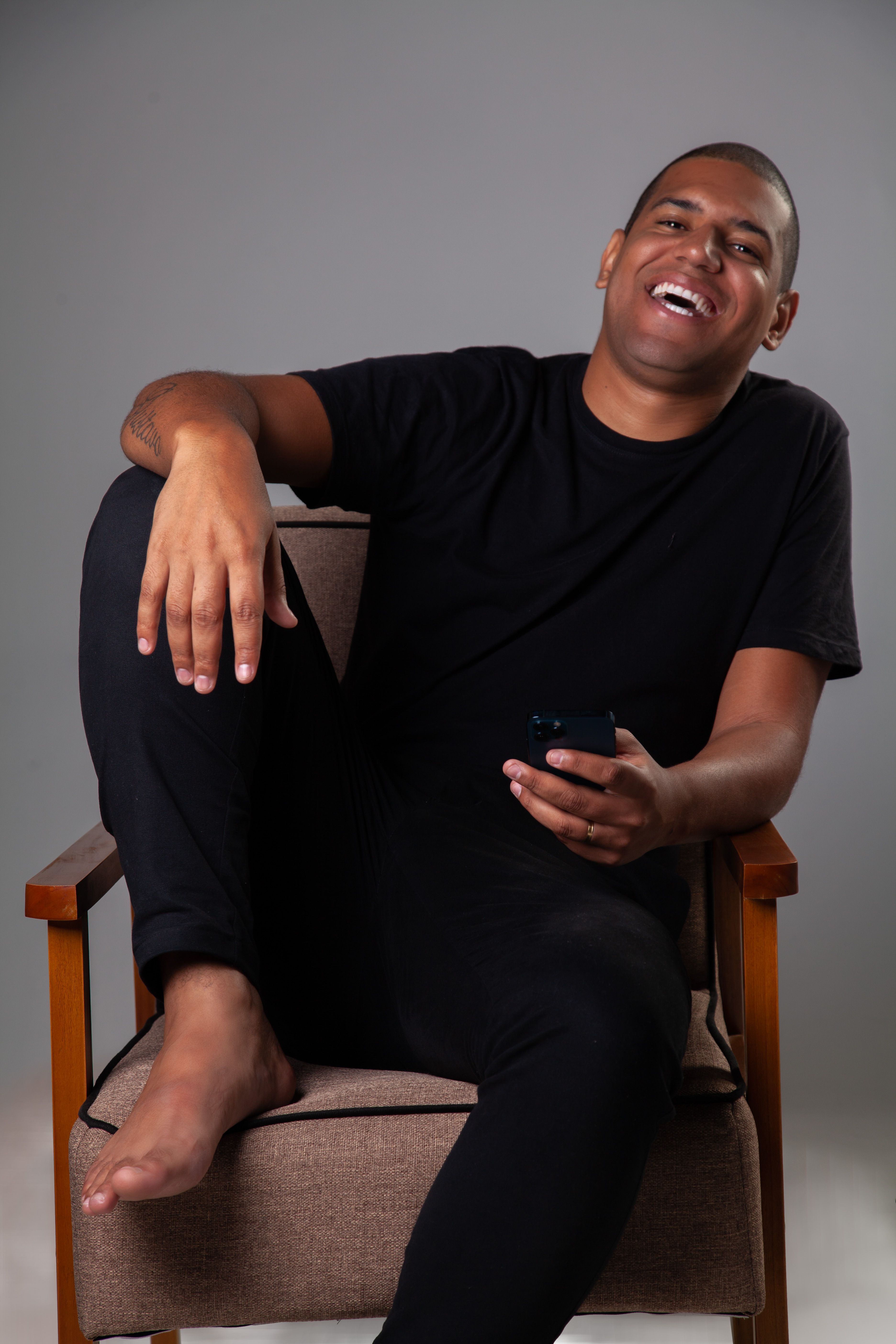 Man in black attire, barefoot, seated on a chair holding a smartphone. Captured indoors.