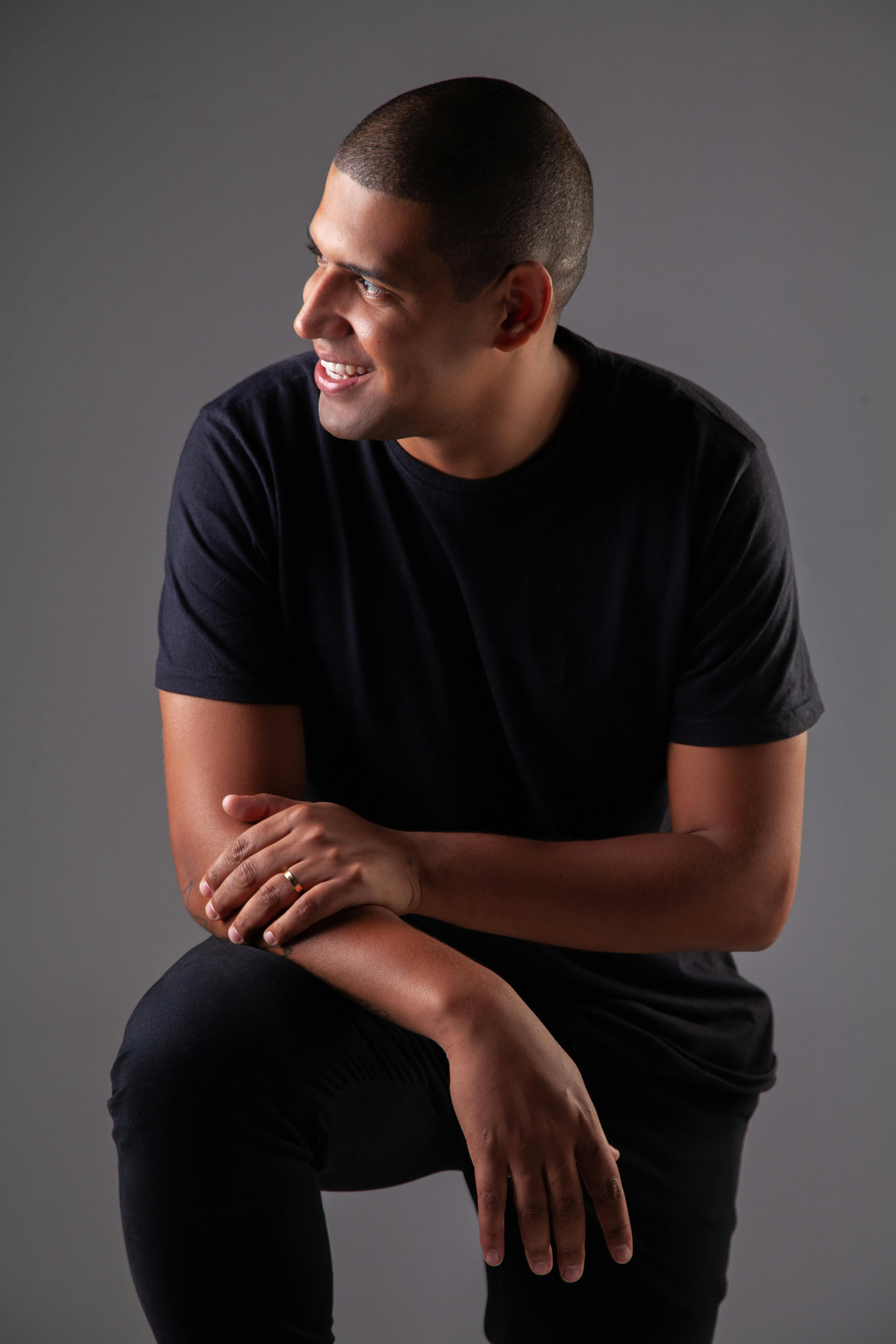 Stylish adult man smiling indoors in a studio setting, wearing black clothes with a gray background.