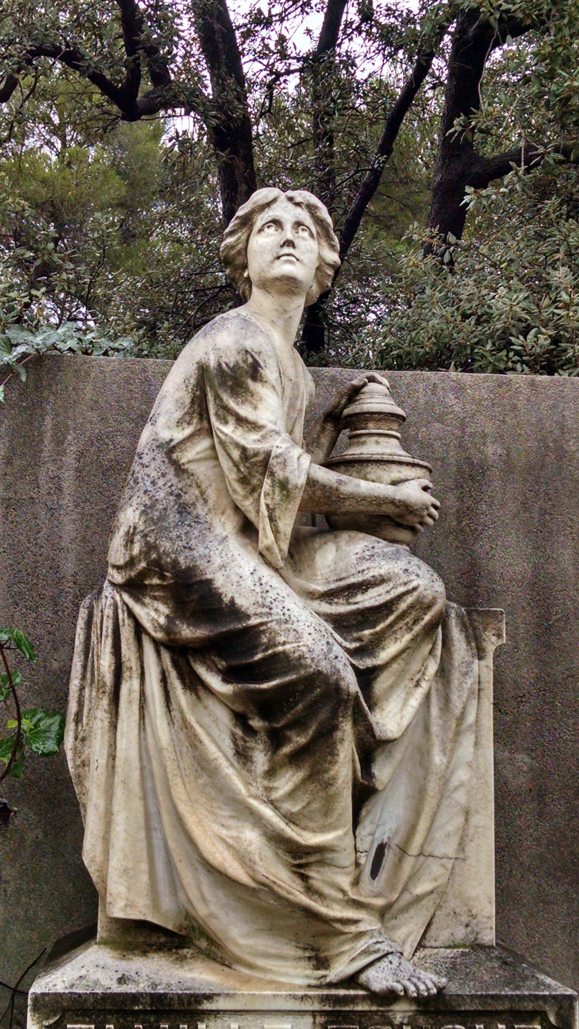 A detailed statue of a woman holding a jar set against lush greenery.