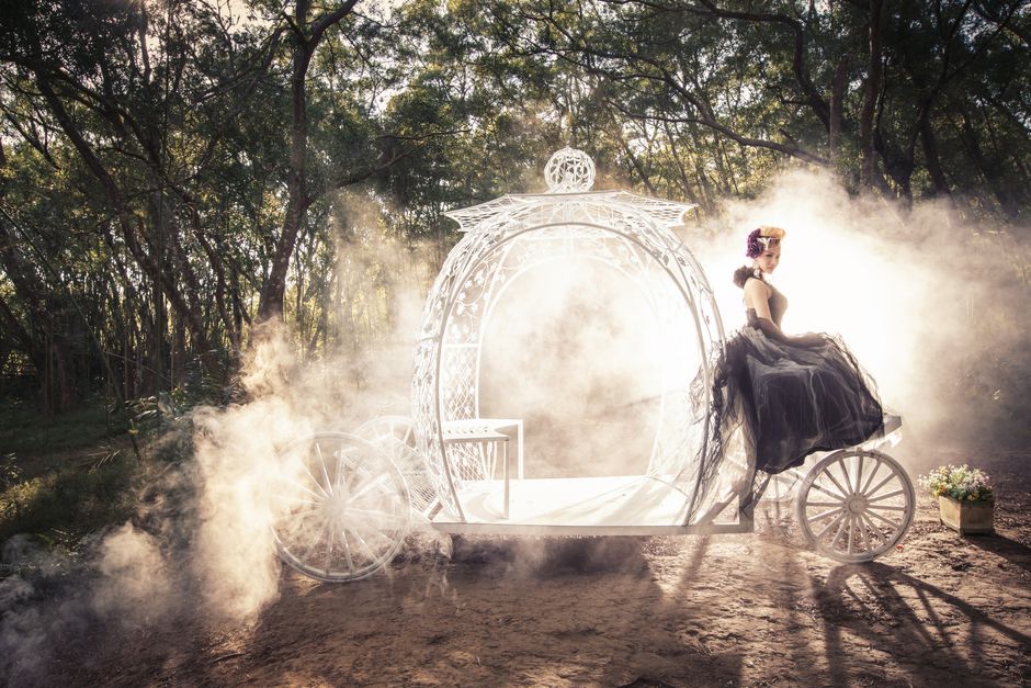 A woman in a vintage gown sits on a mystical carriage in a foggy forest setting.