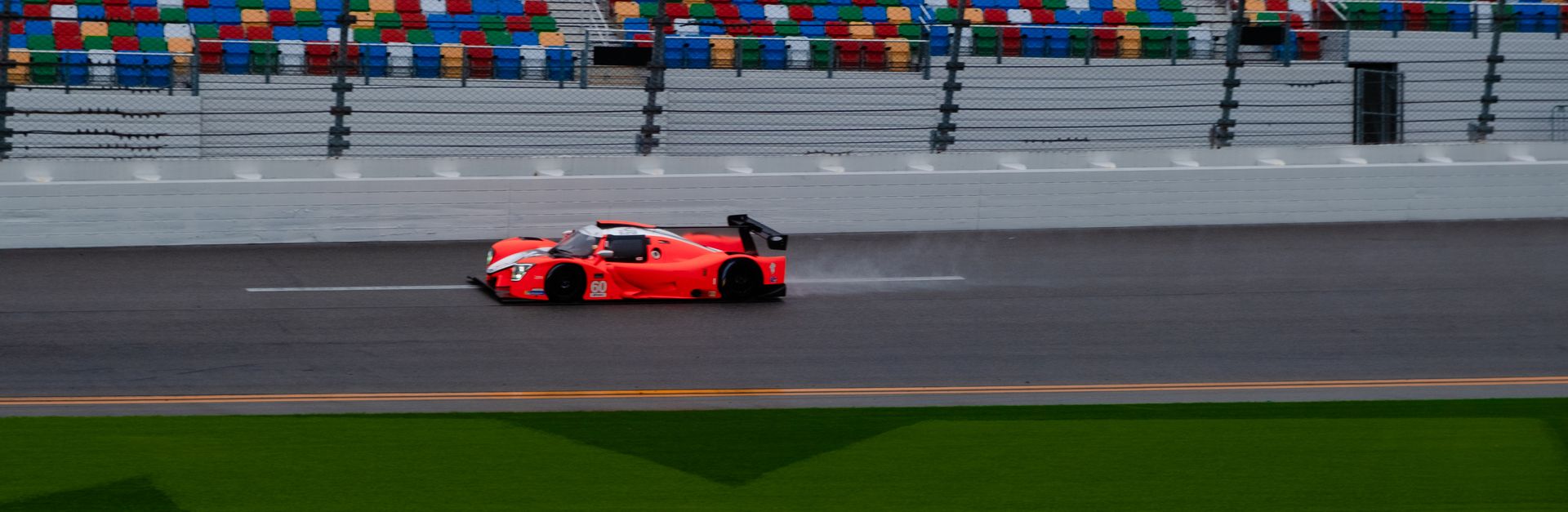High-speed red race car zooms past colorful seats on a rain-slicked track during the day.