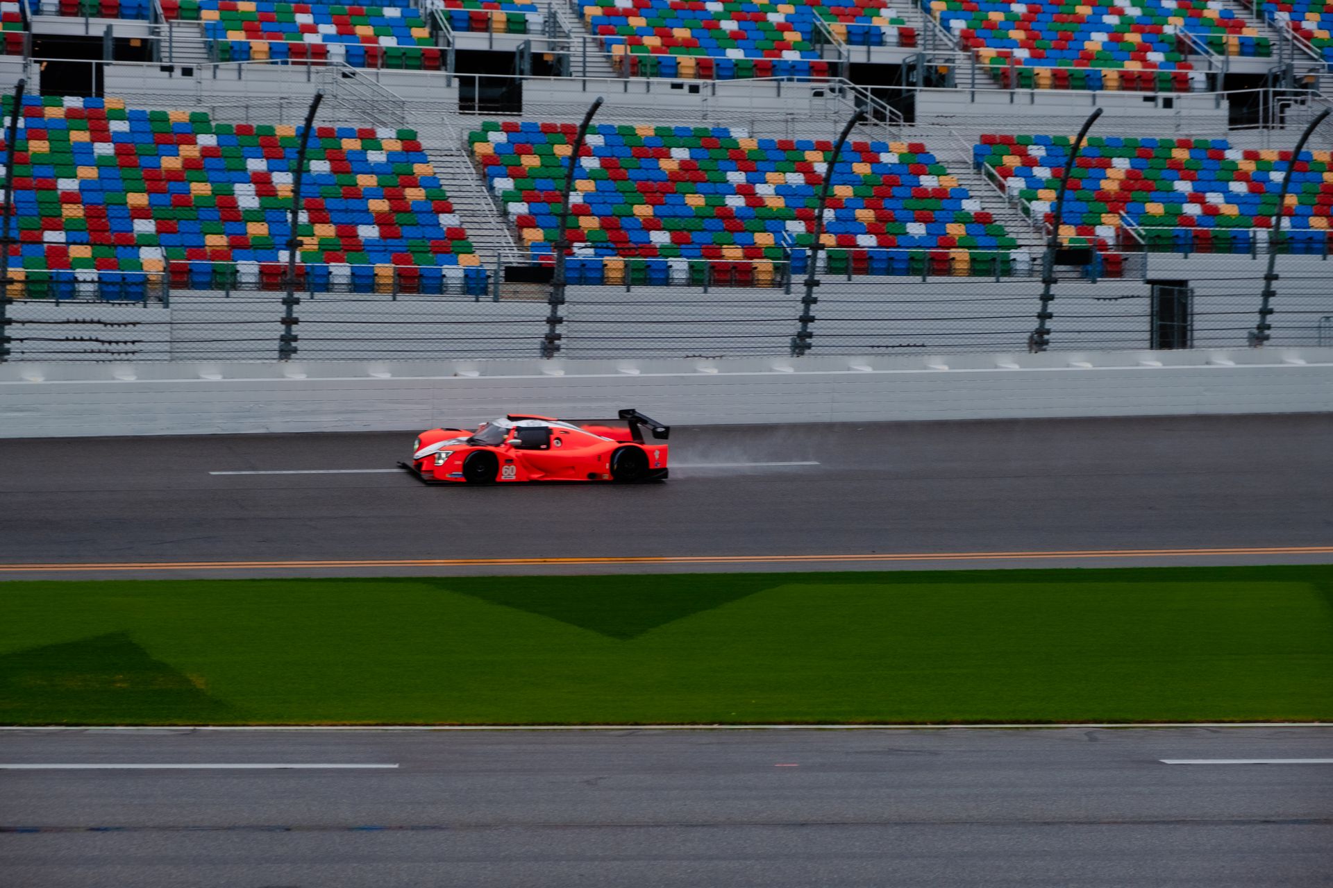 High-speed red race car zooms past colorful seats on a rain-slicked track during the day.
