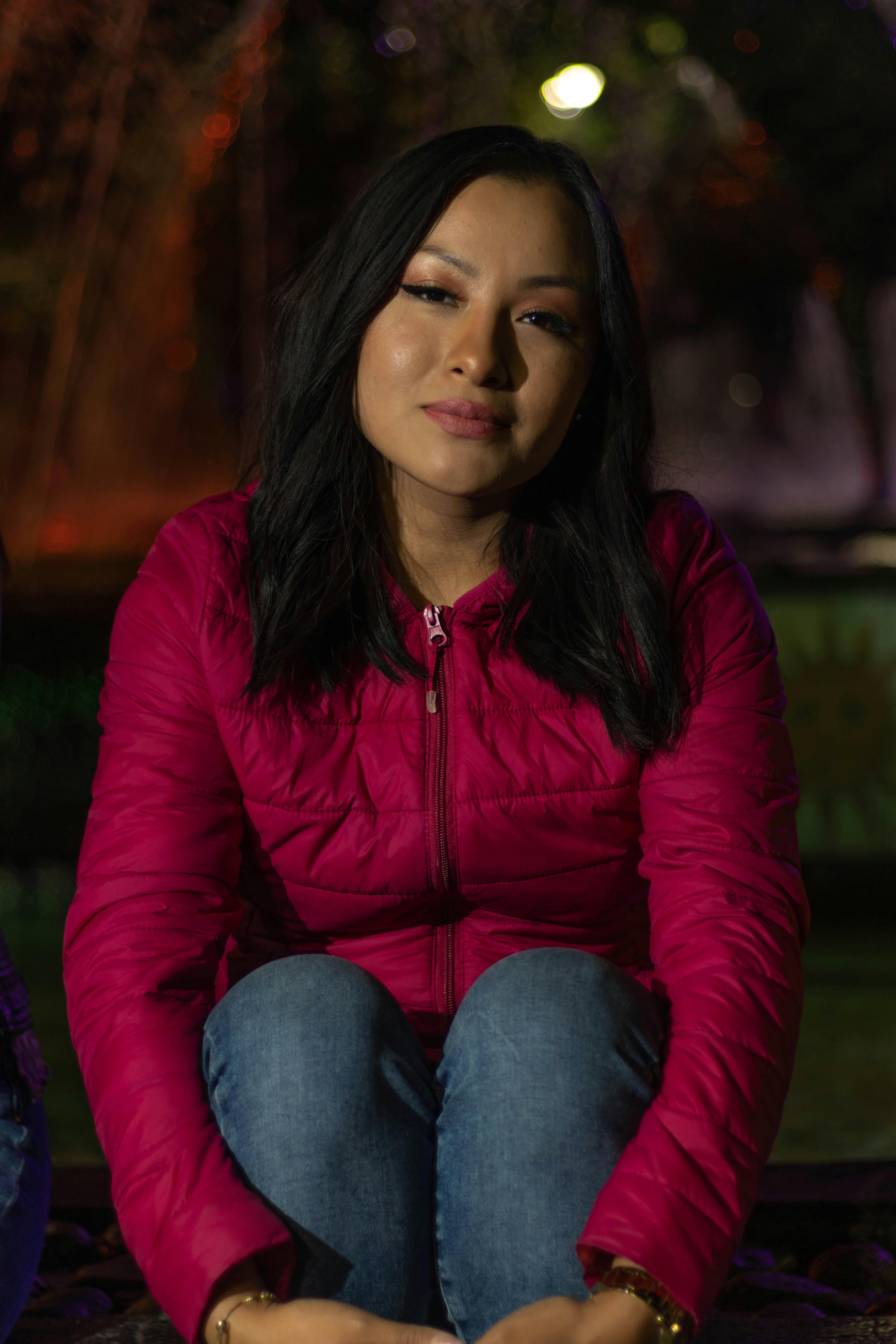 Young woman in a pink jacket sitting outside at night with a thoughtful expression.