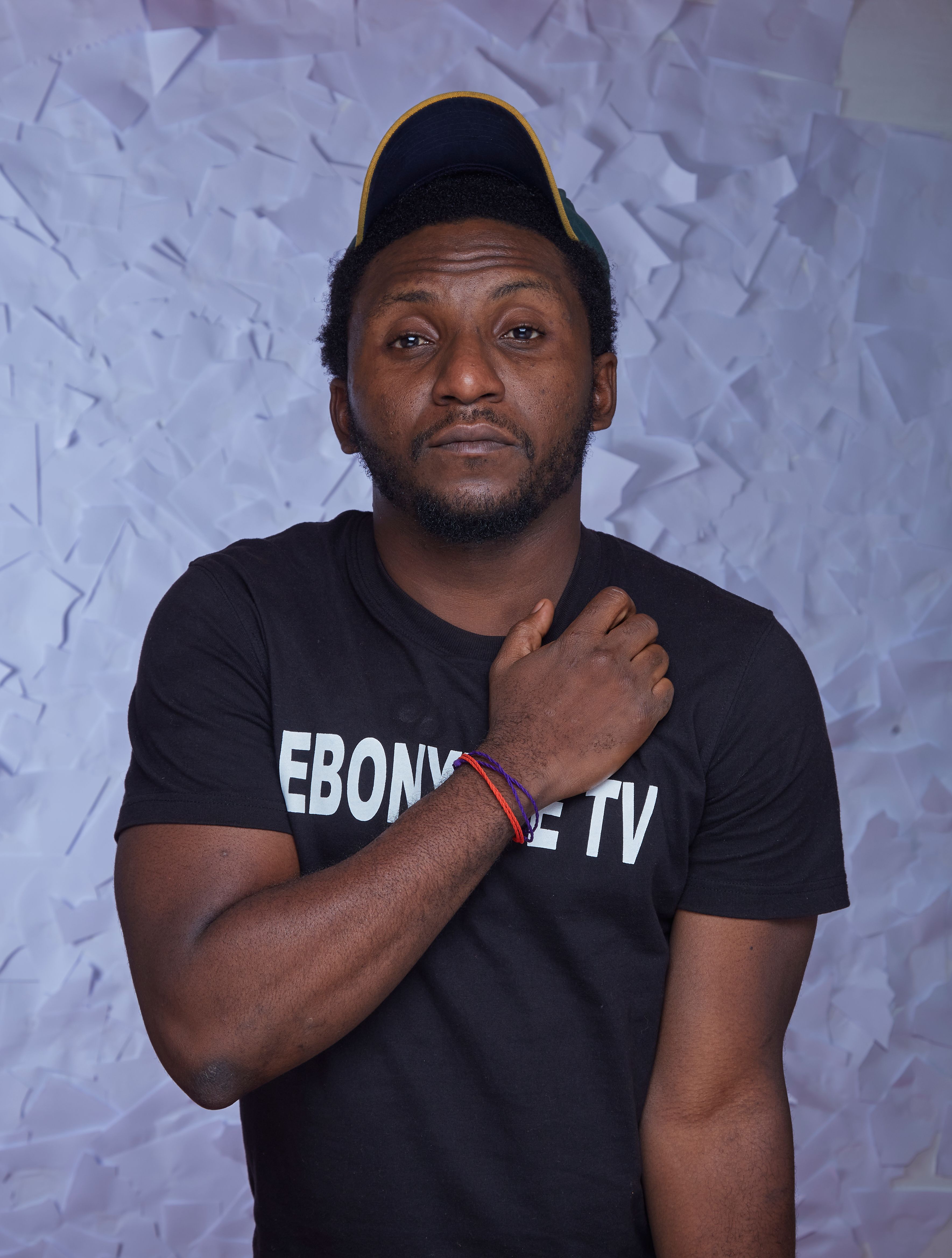 Portrait of a man in a black t-shirt and cap in a studio setting against a textured backdrop.