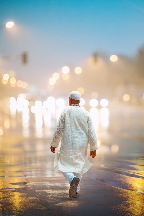 Free Man in traditional attire walks on wet streets of Dhaka at night, with illuminated bokeh background. Stock Photo