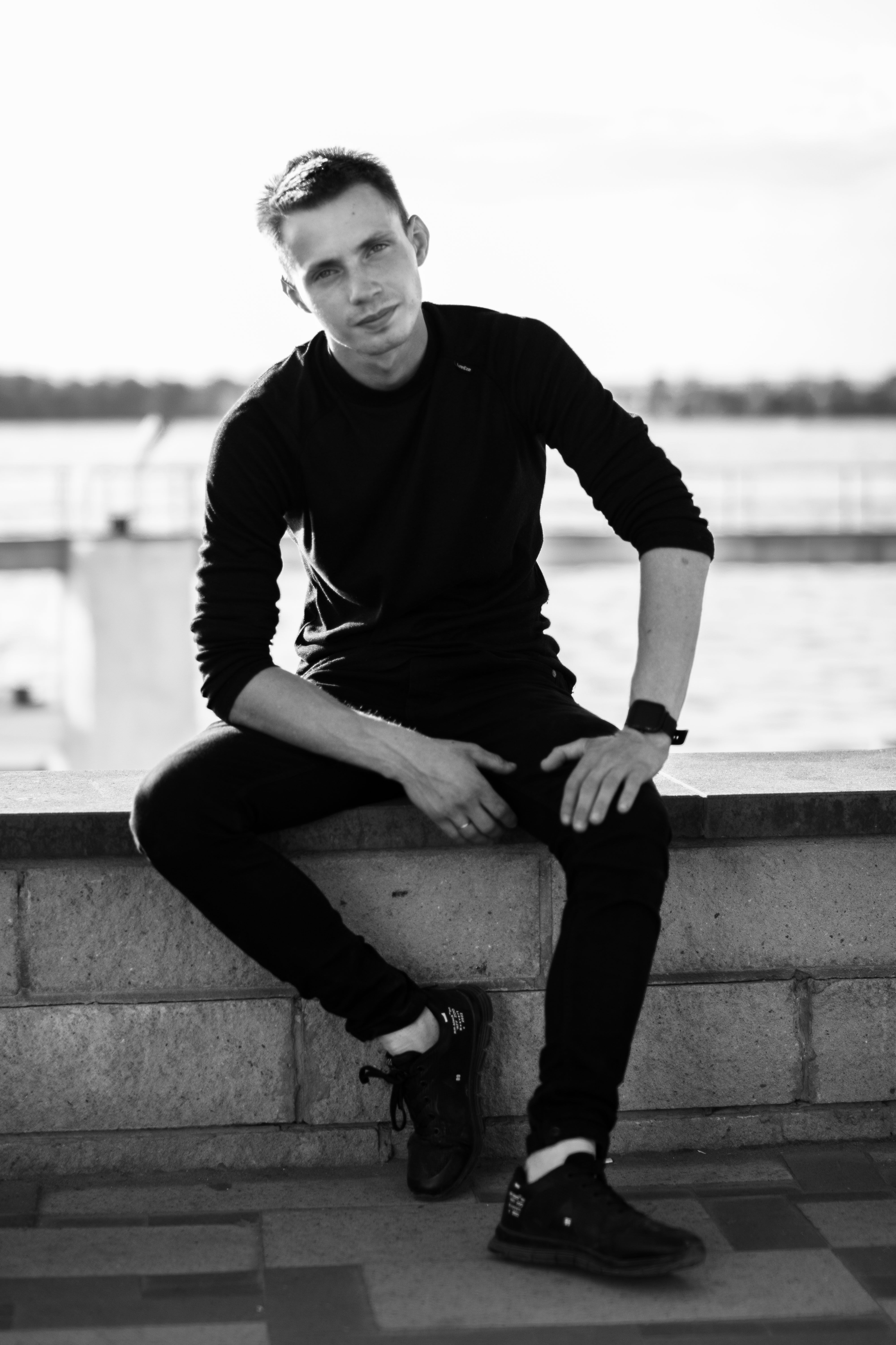 Stylish black and white portrait of a young man sitting by the waterfront, wearing casual attire.