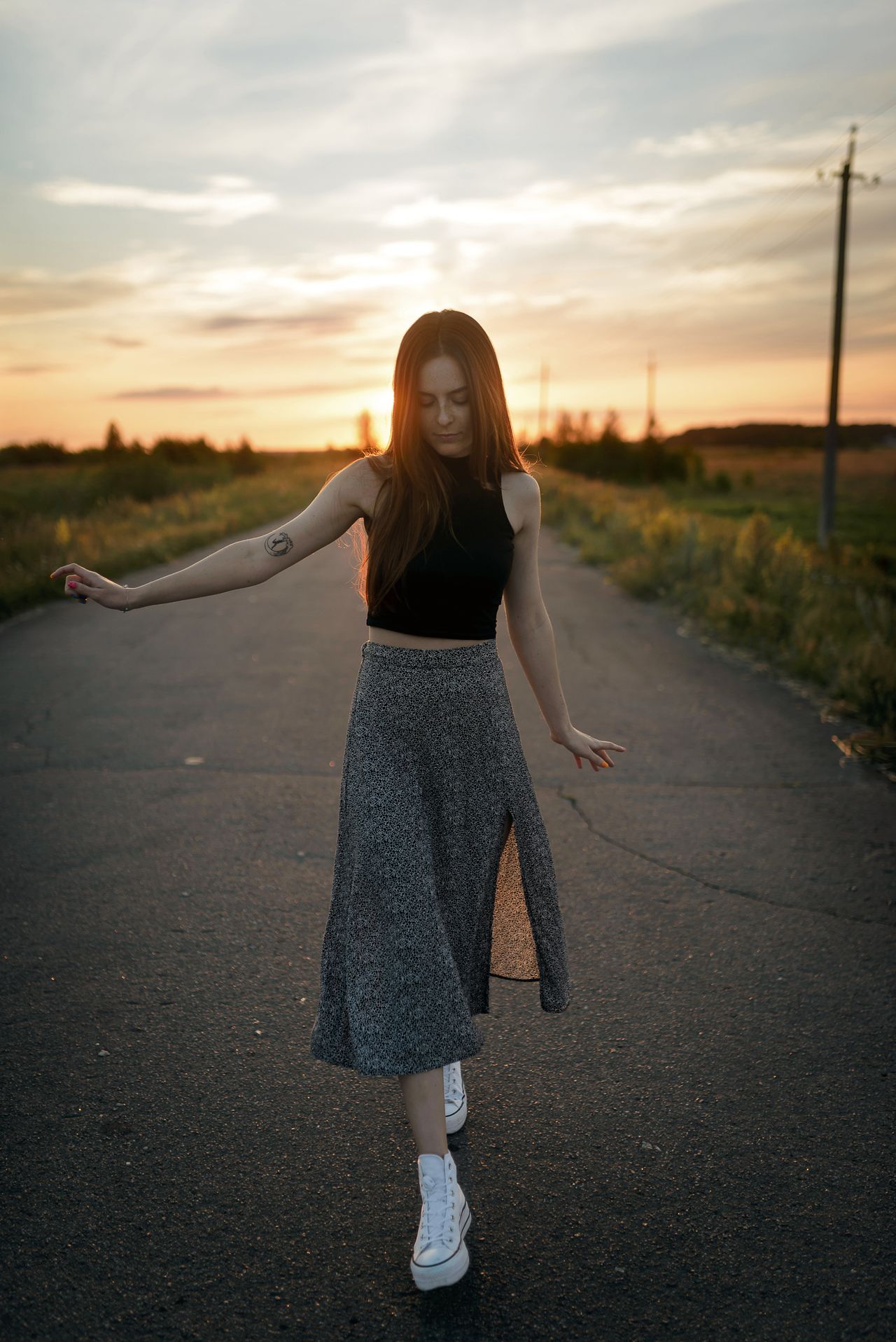 Woman in a black crop top, patterned midi skirt and white sneakers at sunset