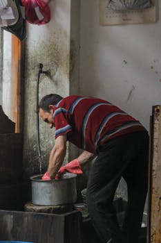 Elderly man in gloves cleaning a metal pot inside an old-style kitchen with a water hose.