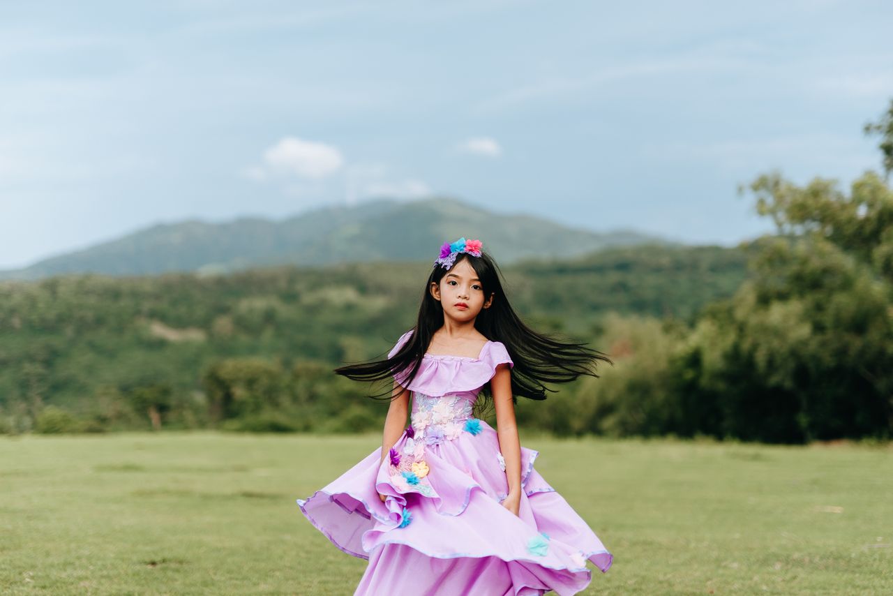a young girl in a vibrant purple dress stands outdoors in a scenic lan