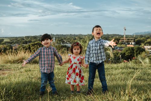 Free Three joyful children holding hands in a grassy field with a scenic background in La Dorada, Colombia. Stock Photo