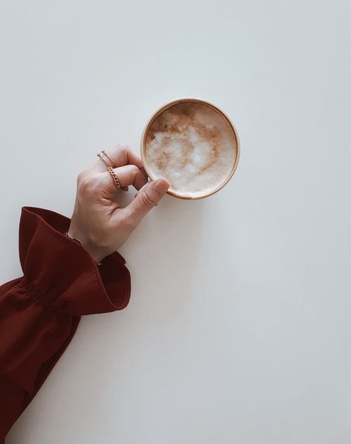 Free Flat lay of a hand holding a cappuccino with frothy latte art on a white surface. Stock Photo