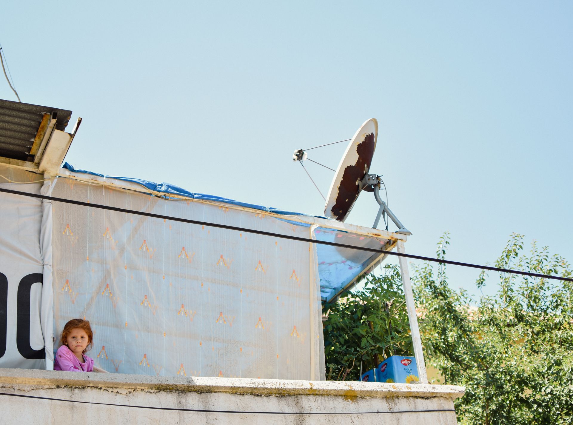 A young child stands on a balcony under a clear sky, next to a satellite dish.