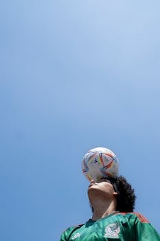 A soccer player showcases balancing skills with a ball under a clear blue sky, highlighting athletic ability and focus.