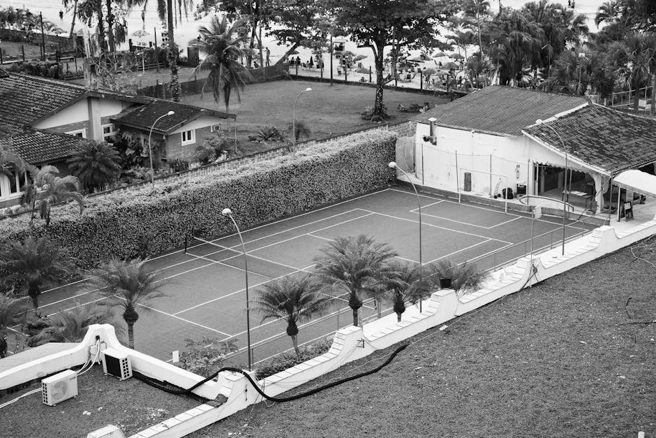 Black and white aerial shot of a tennis court surrounded by tropical plants and buildings.