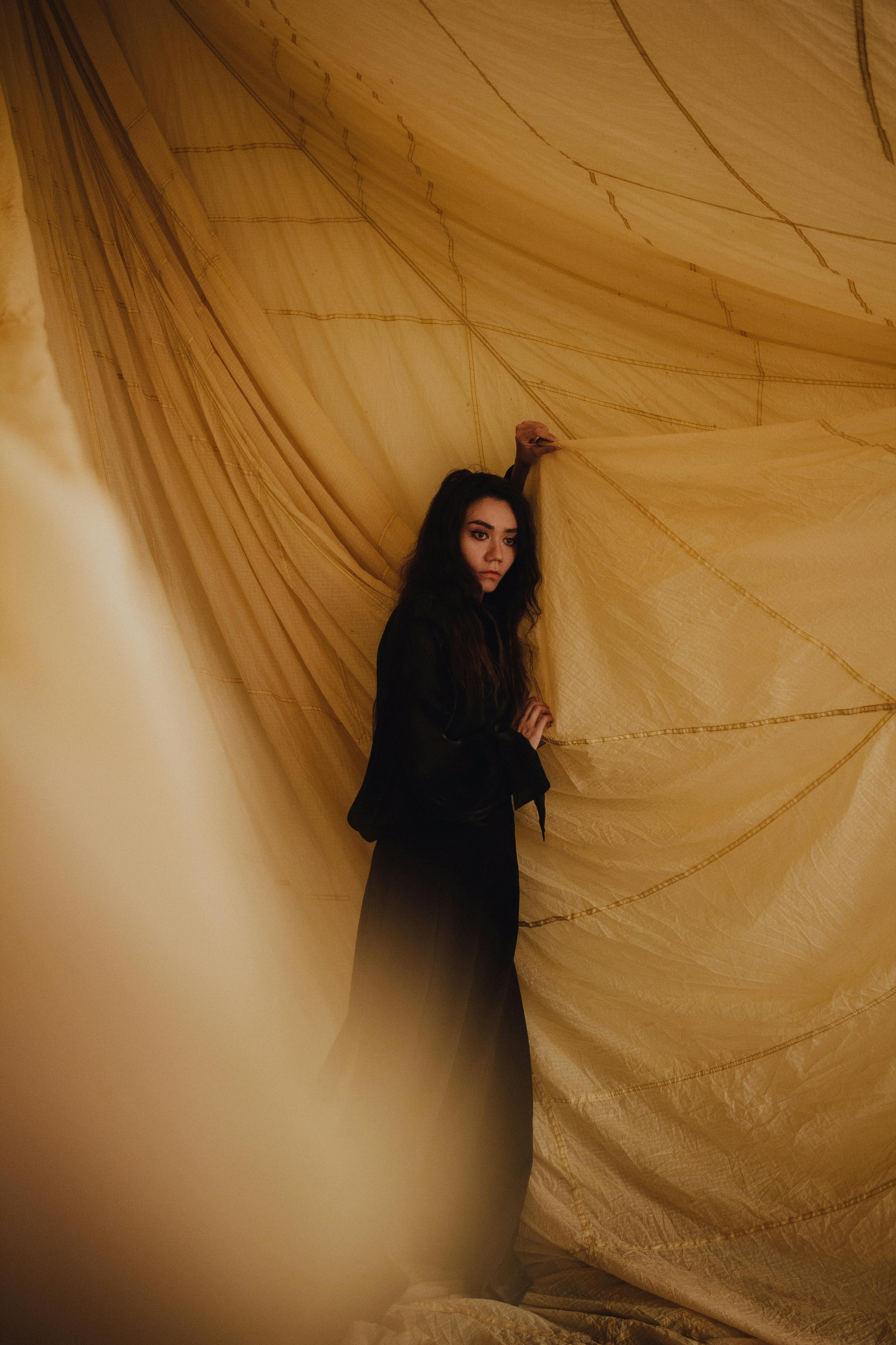 A woman poses indoors with flowing fabric in a warm, dramatic setting.